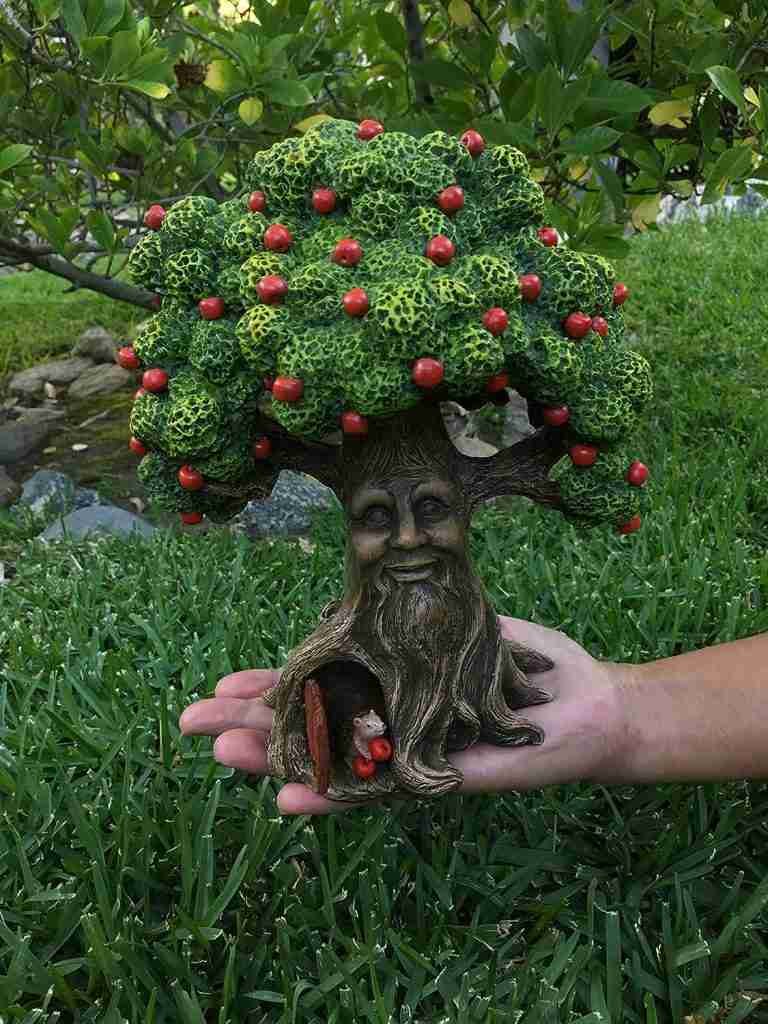 Hand holding a small, decorative tree figurine with a carved face and red berries, showing a hollow with a small animal inside, against a green grassy background with shrubs.