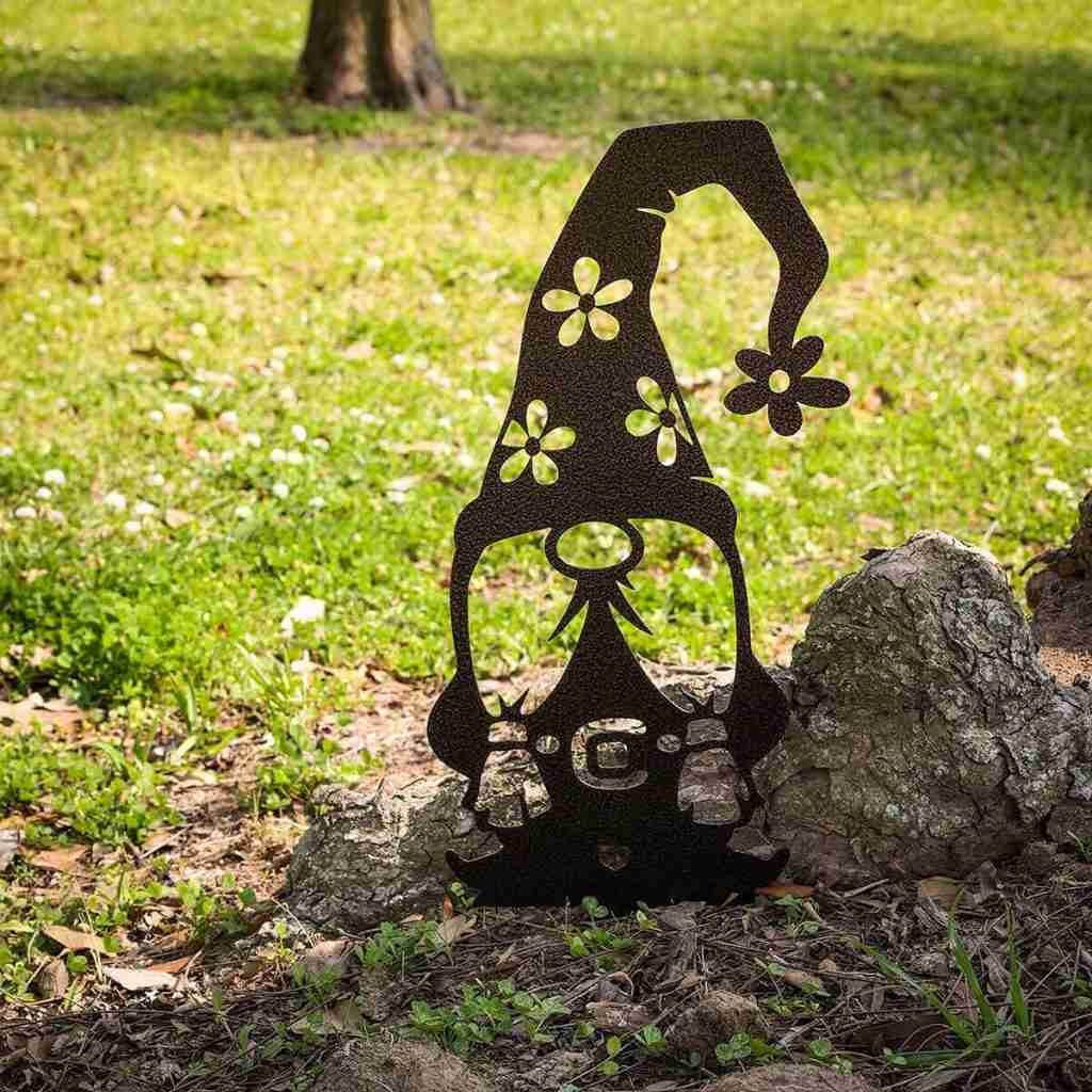Metal garden ornament of a gnome with a tall flower-adorned hat, placed on the ground near a rock in a grassy area.