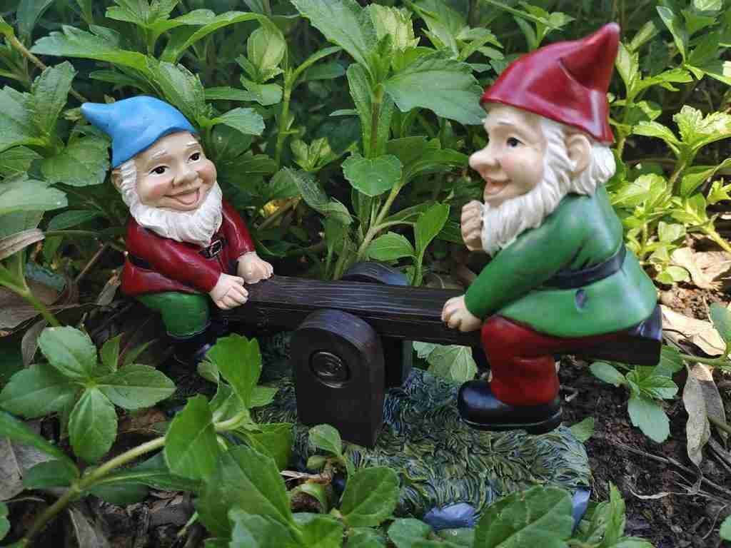 Two garden gnome figurines on a seesaw, surrounded by green foliage.