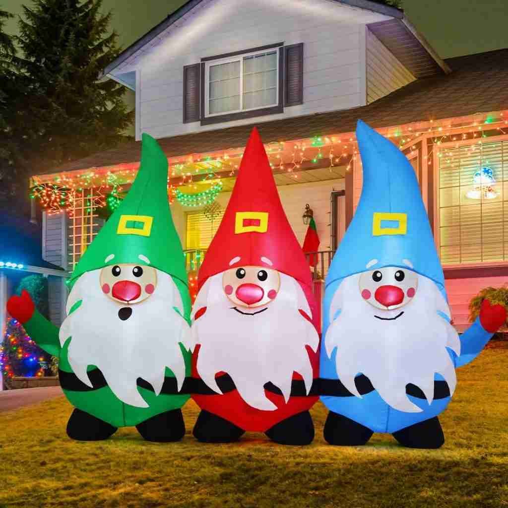 Three inflatable Christmas gnomes with colorful hats and beards in front of a house decorated with Christmas lights.