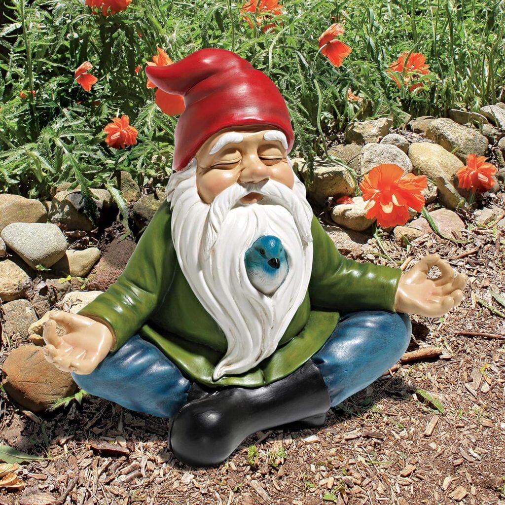 A garden gnome statue wearing a red hat and green coat sitting in a meditative pose among red flowers and rocks.