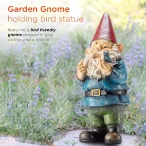 Garden gnome holding a bird statue, standing in a field of purple flowers. Gnome is dressed in blue clothes and a red hat.